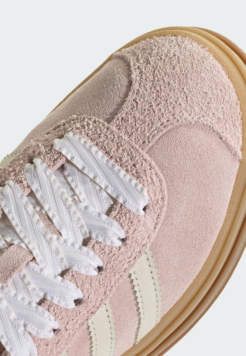 Roze suède sneaker met witte, getextureerde veters, voorzien van drie horizontale witte strepen, een ronde neus en een rubberen zool.