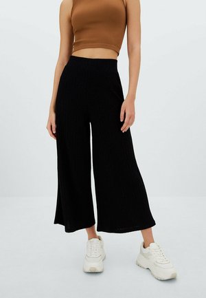 Frau trägt ein braunes ärmelloses Crop-Top, weite schwarze Hosen und weiße Sneakers, steht vor einem schlichten hellen Hintergrund.