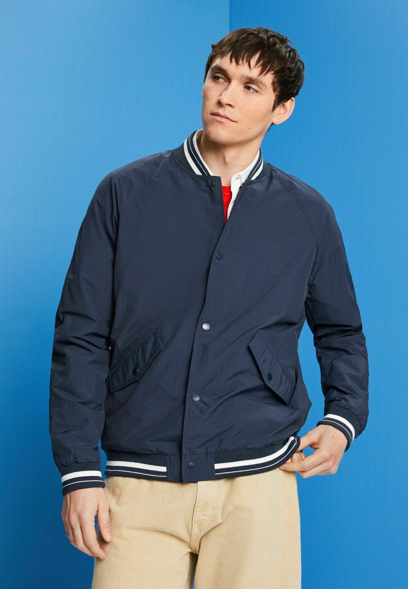 Veste aviateur navy avec des poignets et un col à rayures blanches, dotée de boutons-pression et de poches latérales. Texture lisse, tissu léger.