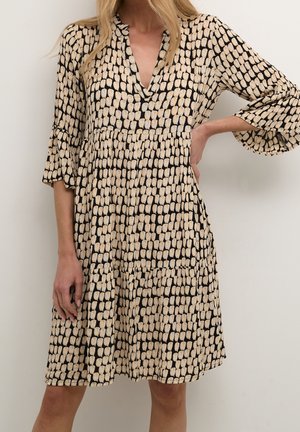 Vrouw draagt een jurk tot op de knie met een beige en zwart abstract bloembladpatroon en driekwart mouwen, hand op de heup.