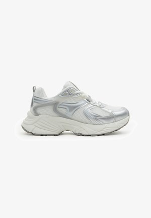 Zapatilla deportiva blanca y plateada con parte superior de malla, suela gruesa y acolchada, cordones y tira de sujeción en el talón, mostrada en perfil lateral.