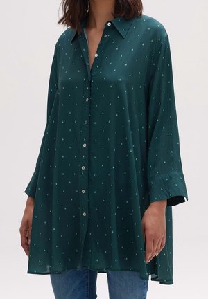 Vrouw met een losse donkergroene blouse met kleine lichtgroene zeilbootjespatronen en een blauwe spijkerbroek, staand tegen een eenvoudige achtergrond.