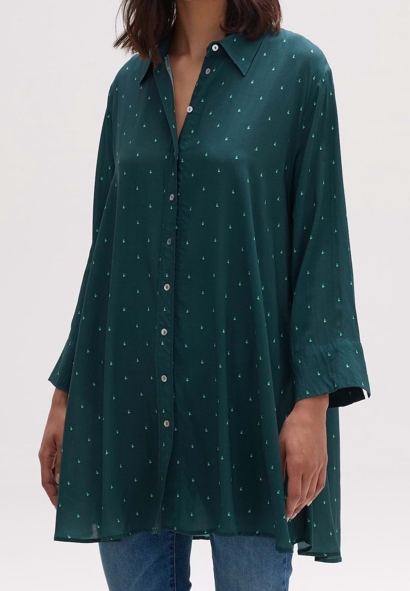 Femme portant une chemise ample vert foncé boutonnée avec de petits motifs de voiliers vert clair et un jean bleu, debout devant un fond uni.