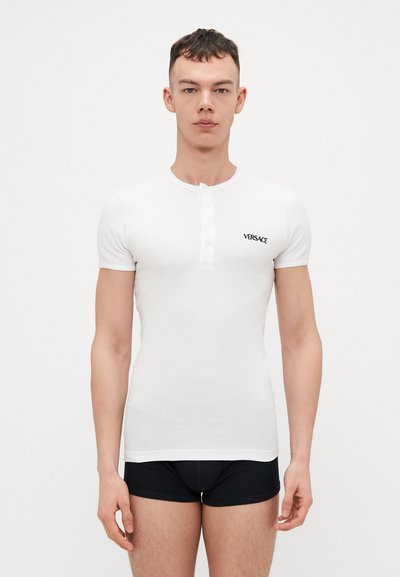 Witte, kortrigelende fitted t-shirt met een knoopsluiting op de hals en een zwart "VERSACE" logo op de linkerkant van de borst, gemaakt van gladde stof.