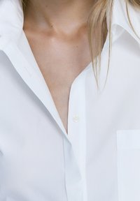 Camicia bianca con colletto classico, dotata di taschino sul petto e texture liscia. I bottoni sono rotondi e realizzati in un materiale chiaro.