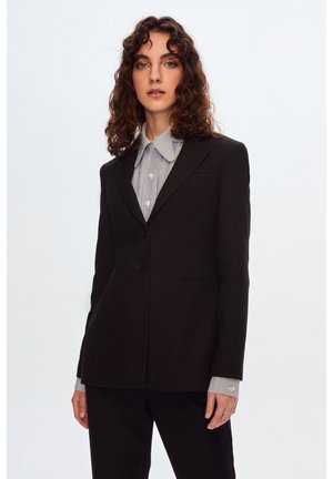 Blazer nero su misura con un unico bottone, rever a punte e due tasche frontali, indossato sopra una camicia a righe con colletto. Texture liscia.