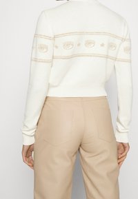 Personne portant un pull court blanc avec des motifs d’œil et d’étoile, associé à un pantalon beige taille haute, vue de dos.