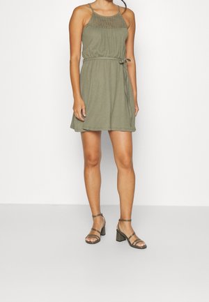 Robe verte, longueur genou, avec un corsage texturé, des bretelles spaghetti et une taille nouée. Associée à des sandales à talons blocs et brides.