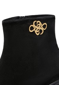 Stivale alla caviglia in suede nero con un emblema decorativo in metallo dorato che presenta un design astratto di anelli intrecciati vicino al bordo superiore.