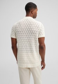 Cremefarbener Kurzarm-Polo mit gestricktem Muster und geripptem Kragen sowie Ärmeln. Getragen mit hellen Hosen.