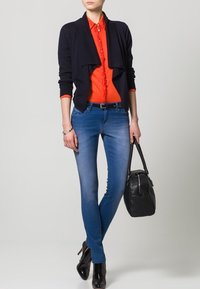 Cardigan aperto di colore blu navy sopra una camicia rossa con bottoni, abbinato a jeans skinny blu chiaro e stivali neri con tacco alto, mentre tiene una borsa nera.