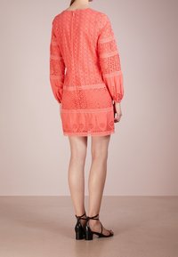 Minišaty korálové barvy s výřezy, dlouhými balónovými rukávy, složitými krajkovými detaily a zaobleným výstřihem na zádech. Kombinovány s černými páskovými podpatky.