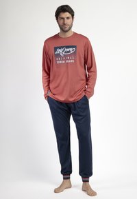 Camisa de manga larga de color óxido con un logotipo gráfico azul, combinada con pantalones deportivos navy con puños a rayas y bolsillos.