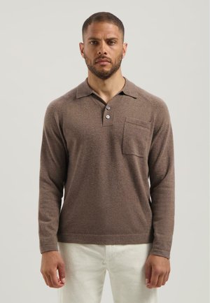 Man in een bruin poloshirt met lange mouwen, voorzien van knopen en een borstzak, gecombineerd met lichte broek, staande voor een effen achtergrond.