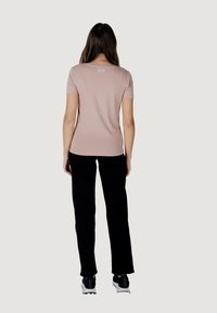 Camiseta de manga corta rosa claro con cuello redondo y un logo sutil en la parte posterior, combinada con pantalones largos negros y zapatillas deportivas negras.