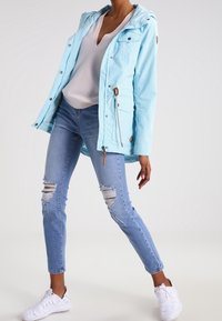 Ljusblå, vattentät jacka med huva och framfickor, kombinerad med slitna blå jeans och vita sneakers.