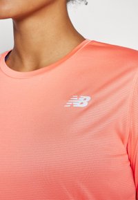 Korte mouwen, koraalkleurig sportshirt van gemarkeerde stof. Heeft platte naden en een wit "New Balance" logo op de borst.
