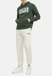 Donkergroene hoodie met witte "JACK & JONES" graphic, gecombineerd met lichtgrijze joggers met logo. Witte sneakers maken de outfit compleet.