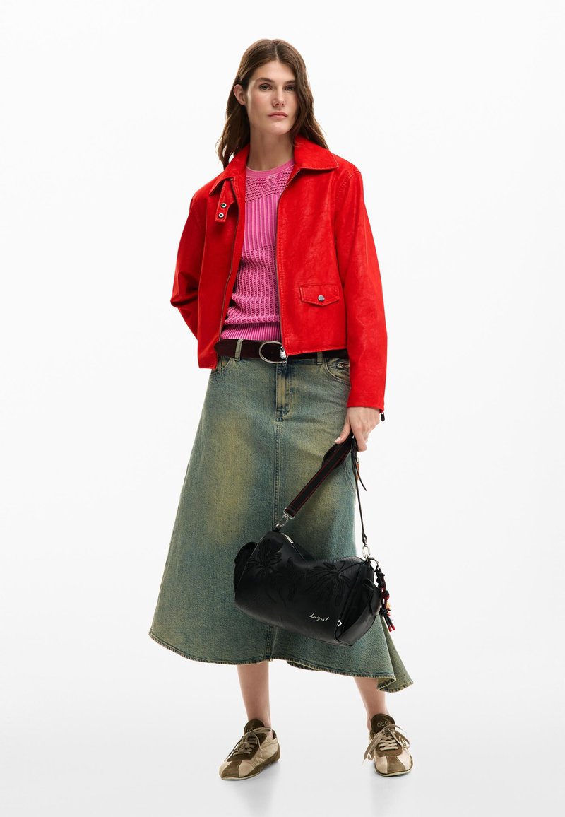 Mujer vistiendo una chaqueta roja, top rosa, falda vaquera verde, zapatillas beige, sosteniendo un bolso negro con diseños florales.