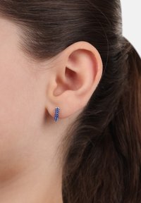 Pendiente en forma de aro de cristal azul con un diseño elegante y redondeado. Adornado con múltiples pequeñas piedras azules, creando una apariencia texturada.