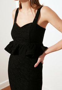 Abito in pizzo nero con spalline larghe, un busto aderente e una ruffles a peplum in vita, con una texture liscia e motivi intricati.