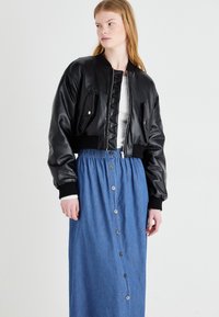 Giacca bomber nera in pelle, tagliata, con parte frontale trapuntata, tasche laterali e polsini a coste, indossata su una gonna in denim azzurro con bottoni.