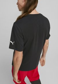 Camiseta de manga corta negra hecha de algodón, con cuello redondo y un logo blanco de Puma en la manga izquierda. Textura suave, ajuste relajado.