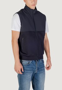 Gilet nero senza maniche con collo alto, chiusura con zip, tasche laterali e una texture liscia; abbinato a jeans in denim azzurri.