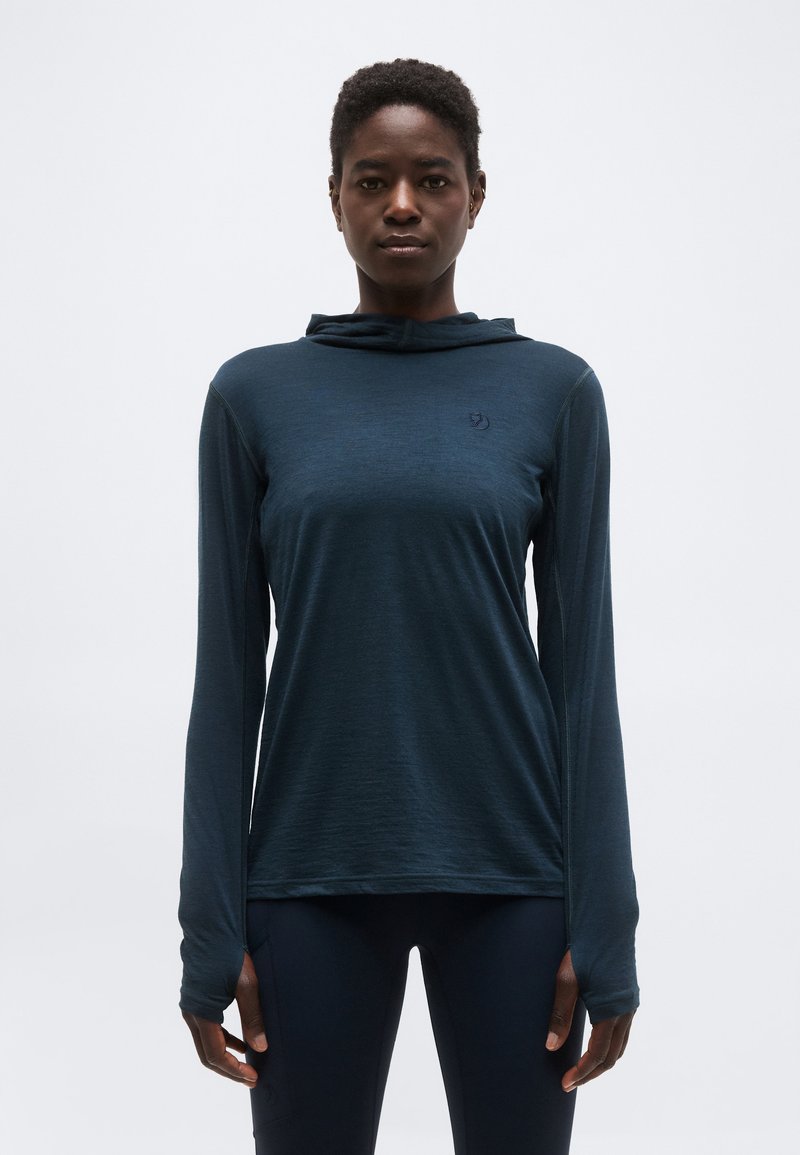 Woman wearing a dark blue long-sleeve hoodie with thumb holes, standing against a plain light background facing forward.