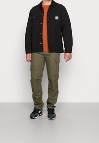 Czarna kurtka z płótna z dwoma kieszeniami na piersi, pomarańczowy sweter, oliwkowe spodnie cargo i czarne trampki z białymi akcentami. Gładkie tekstury.