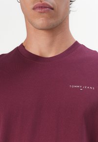 Burgunderfarget bomull t-skjorte med rund hals, med en liten hvit "TOMMY JEANS" logo på venstre brystområde. Glatt tekstur, normal passform.