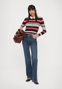 Randig stickad tröja i nyanser av beige, rött och svart, kombinerad med blå utsvängda jeans. Håller en brun handväska.