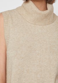 Kvinna iklädd en ärmlös beige stickad turtleneck-tröja som visar den övre delen av torso och hals.