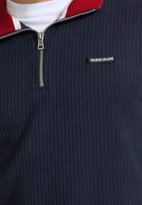 Felpa con zip a costine blu navy con colletto rosso, cerniera argentata e toppa con logo nero "GUESS JEANS" sul petto a sinistra.