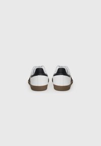White sneakers with black accents, textured black panel, and brown rubber sole. Smooth finish, low-top design, and rounded toe.