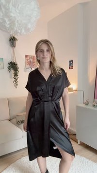 Μαύρο satiny φόρεμα με κοντά μανίκια, V-λαιμόκοψη, ζώνη στη μέση και πλαϊνό σκίσιμο. Λείο υλικό και μήκος μέχρι το γόνατο.