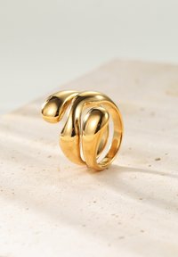 Złoty pierścień o płynnej formie, z trzema wypukłymi, zaokrąglonymi elementami i gładkim wykończeniem, umieszczony na jasnym kamiennym podłożu.