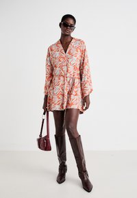 Robe portefeuille florale en orange et bleu clair avec des manches longues, une taille cintrée et un détail à nouer. Assortie à des bottes brunes hauteur genou et un sac bordeaux.