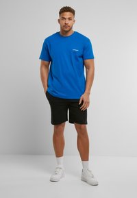 T-shirt en coton bleu avec des manches courtes, un col rond et un petit logo blanc sur la poitrine, associé à un short noir et des baskets blanches.