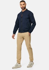 Veste de bombardier navy avec fermeture éclair à l'avant, poignets en côte et poches latérales. Associée à un pantalon beige et des baskets de couleur claire. Design simple et classique.