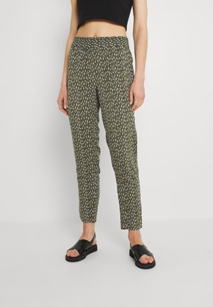 Kobieta nosząca oliwkowozielone, wzorzyste spodnie do kostek, czarny krótki top i czarne sandały z odkrytymi palcami, stoi na białym tle.