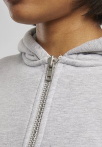 Graue Zip-Hoodie mit weicher Textur, ausgestattet mit einem sichtbaren Metallreißverschluss und einer Kapuze. Der Stoff wirkt leicht und lässig.
