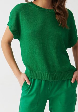 Kobieta ubrana w jaskrawozieloną, krótko rękawową dzianinową bluzkę i dopasowane zielone spodnie, stojąca z rękami w kieszeniach na jednolitym tle.