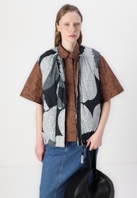 Svart och vitmönstrad väst över en brun skjorta med blommönster, kombinerad med en denimkjol, svart handväska och neutral bakgrund.