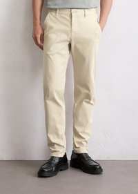 Man in beige chinos en zwarte leren instappers, staande tegen een witte gestructureerde muur met handen in de zakken.