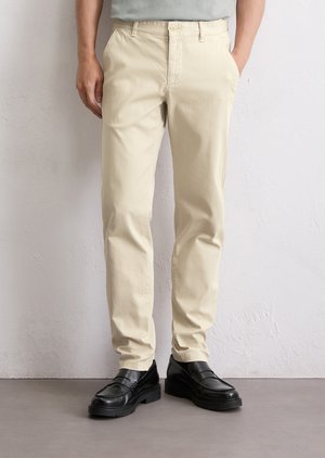 Man in beige chinos en zwarte leren instappers, staande tegen een witte gestructureerde muur met handen in de zakken.