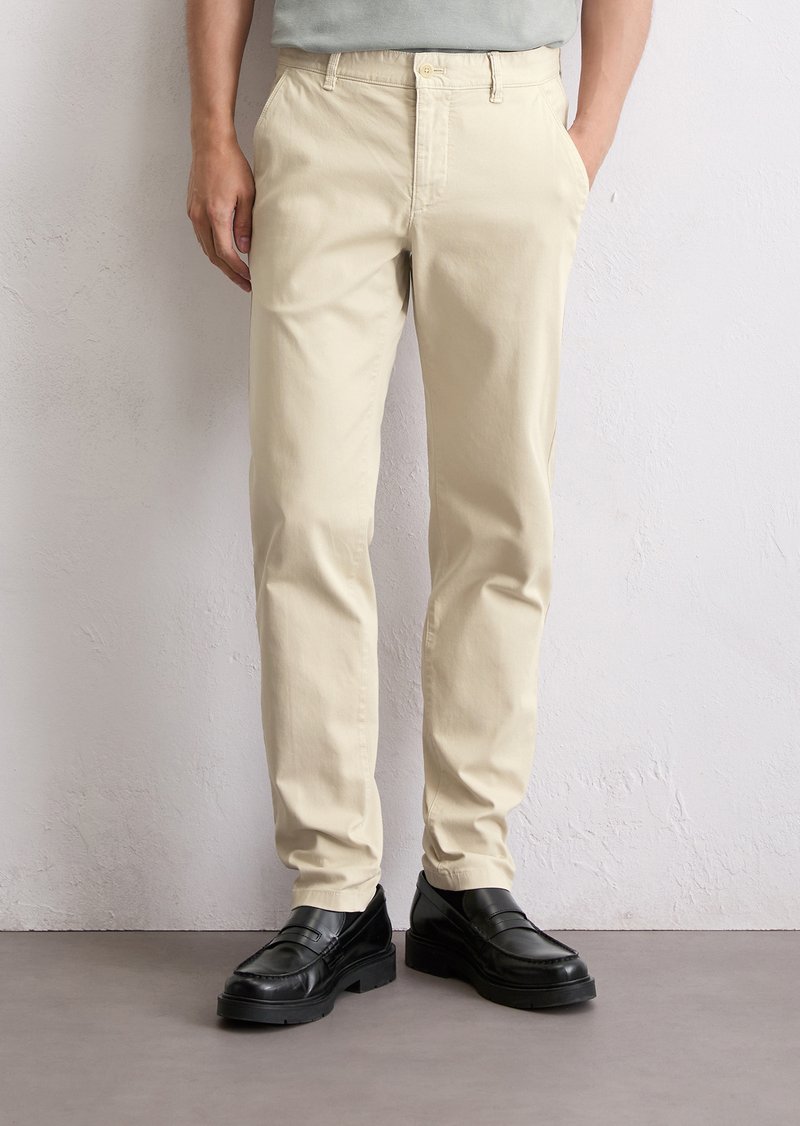 Man in beige chinos en zwarte leren instappers, staande tegen een witte gestructureerde muur met handen in de zakken.