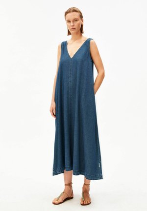 Frau trägt ärmelloses blaues Jeans-Midikleid mit V-Ausschnitt und braunen Riemchensandalen, steht vor einfachem weißem Hintergrund.