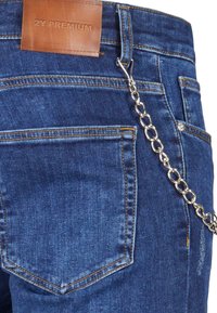 Blå denim jeans med en lærlabel i taljen, udstyret med en sølvkæde, der er fastgjort til bælteløkken, og diskrete gule syninger.