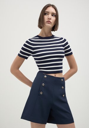 Jeune femme posant avec les mains sur les hanches, portant un crop-top à rayures bleu marine et blanches à manches courtes et un short bleu marine avec des boutons en argent.